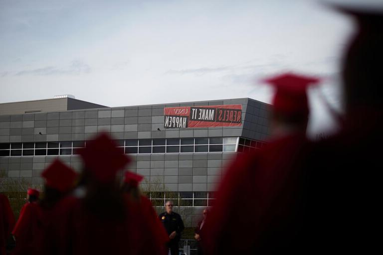 Students in red caps and gowns walk out of an arena in front of a red Rebels Make It Happen sign