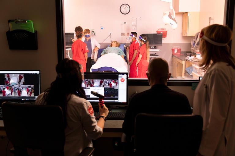 三名临床医生从玻璃后面观察模拟病房中的诊断屏幕和学生. 在观察镜的另一边，学生们倾向于一个人体模型.